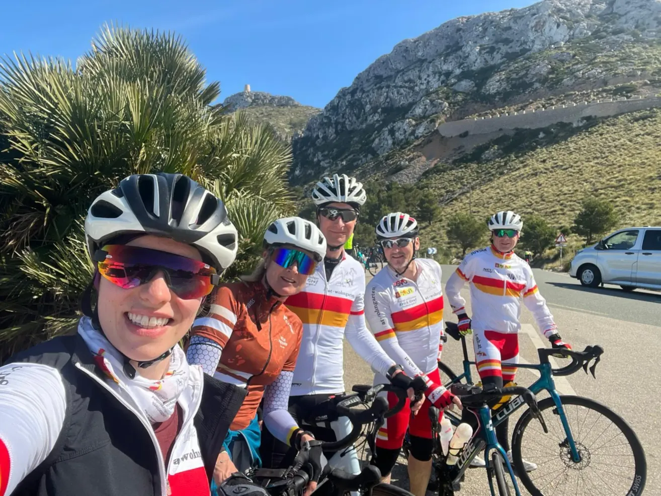 A group of cyclists, all wearing helmets, pose for a photo against a backdrop of mountains and greenery. The cyclists are smiling and appear to be in good spirits.