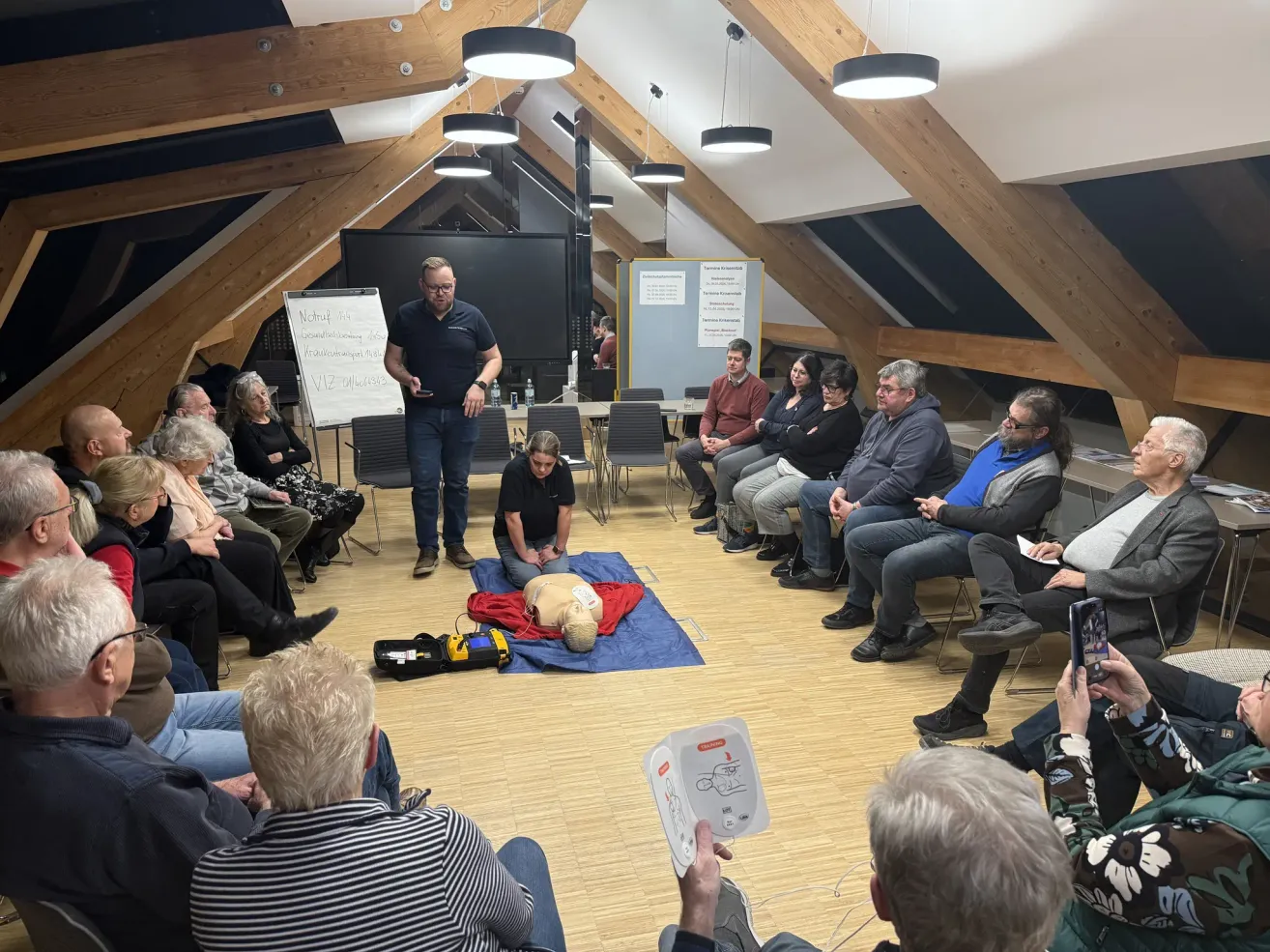 In a room with wooden beams, a man stands by a CPR dummy while others observe. Some people sit in chairs, and one holds a cellphone. A whiteboard and posters are nearby.