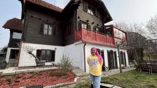 A woman with pink hair stands in front of a house with a red balcony. The house is painted in a combination of white and brown. There is a potted plant and a small table in front of the house.