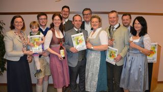 A group of people in traditional German attire pose with awards and wine glasses, smiling for a photo. They hold certificates with event details.