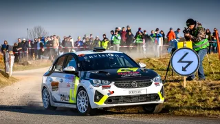 Ein Rallyeauto fährt auf einer Straße, wobei eine Menschenmenge zusieht. Das Auto hat die Nummer 20 auf dem Dach und ist mit Staub bedeckt. Die Menge steht hinter einem Absperrband und trägt Jacken und Mützen.