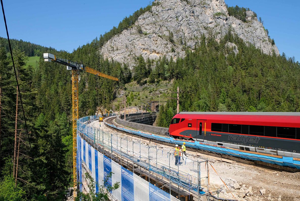 A red train is on the tracks with construction barriers. A crane stands nearby with workers in yellow vests. The train is in front of a rocky hill with trees.