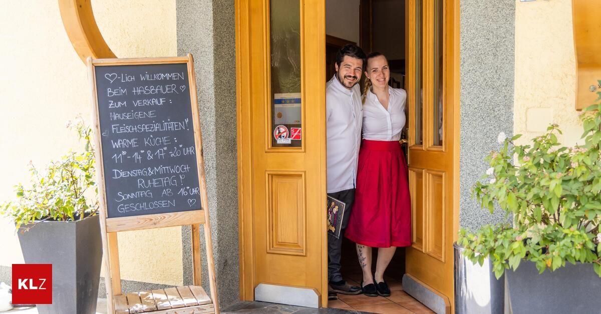 Two people, a man and a woman, are standing in the doorway of a restaurant. They are smiling and facing the camera. A blackboard with text is on the left.