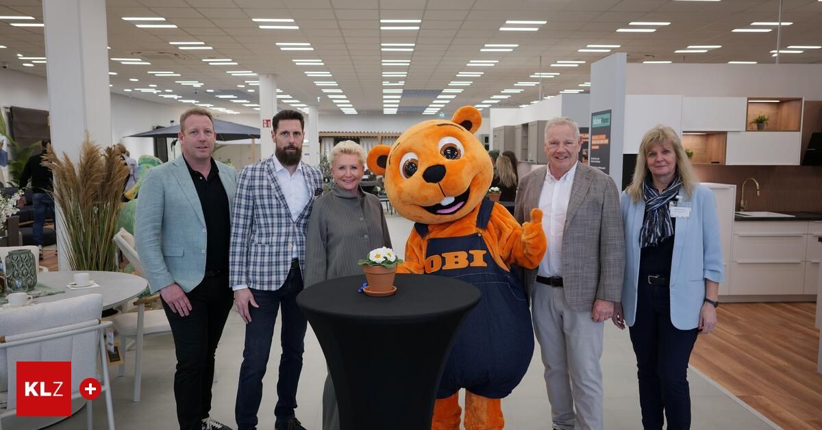Five people are posing for a photo in an indoor area. A mascot is standing in front of a table with a flower pot. Behind them are cabinets and a whiteboard.