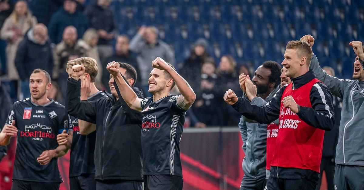 A soccer team celebrates on the field with fans in the stands. The player in the center has his hands raised in triumph.