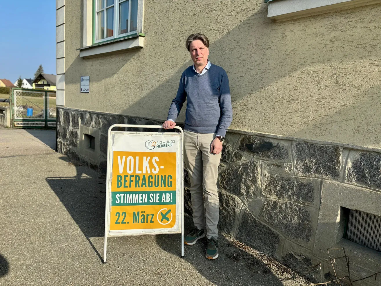 A man stands in front of a building, holding a sign that says 'VOLKS-BEFRUGUNG STIMME SIE AB! 22. März'. The building has a stone wall and a window.
