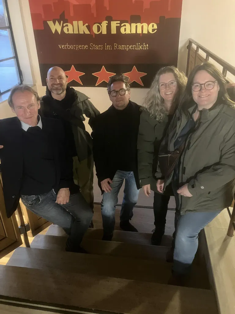 Five individuals stand on a staircase. They all wear winter clothing and glasses. Behind them is a sign with three red stars.