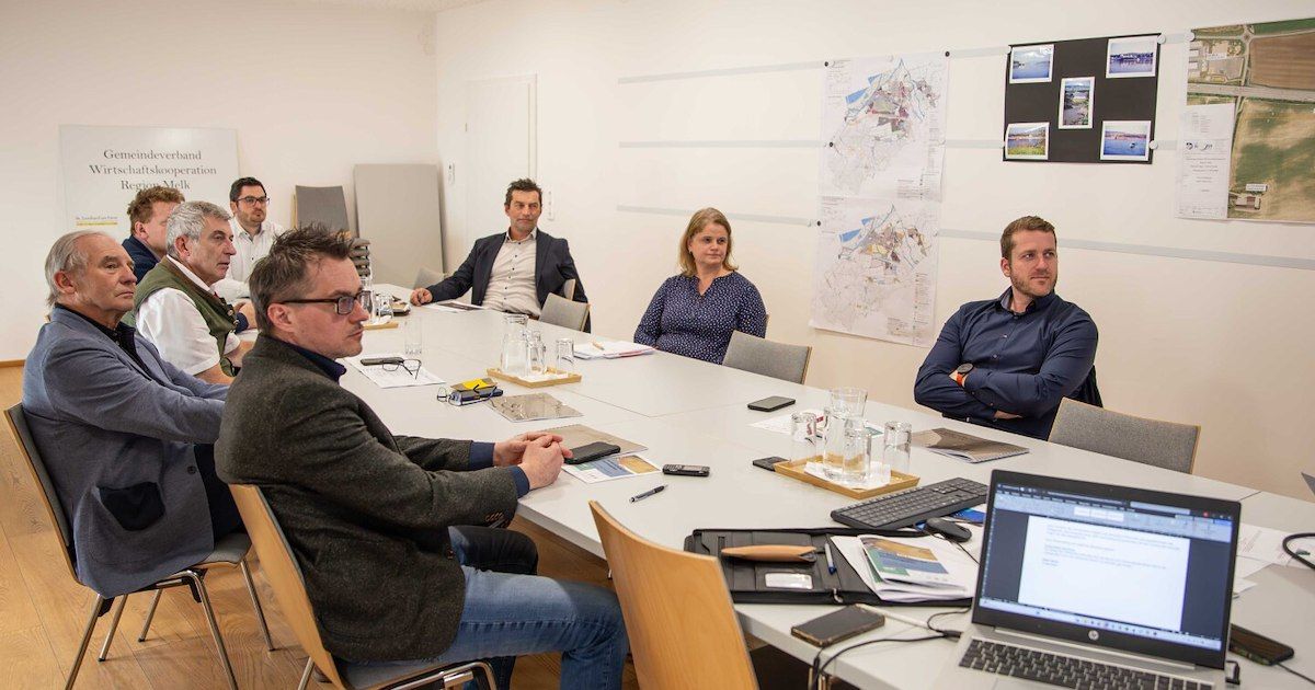Fünf Personen sitzen um einen Konferenztisch herum und sind in einem Meeting verstrickt. Papiere, Laptops und Gläser sind auf dem Tisch. Karten sind an der Wand angeheftet.
