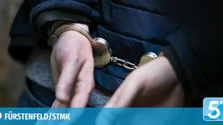 A person is handcuffed, wearing a blue jacket and a black shirt. The handcuffs are connected with a chain, and the person's hands are in front. The image has a watermark with the text 'ELD/STMK'.