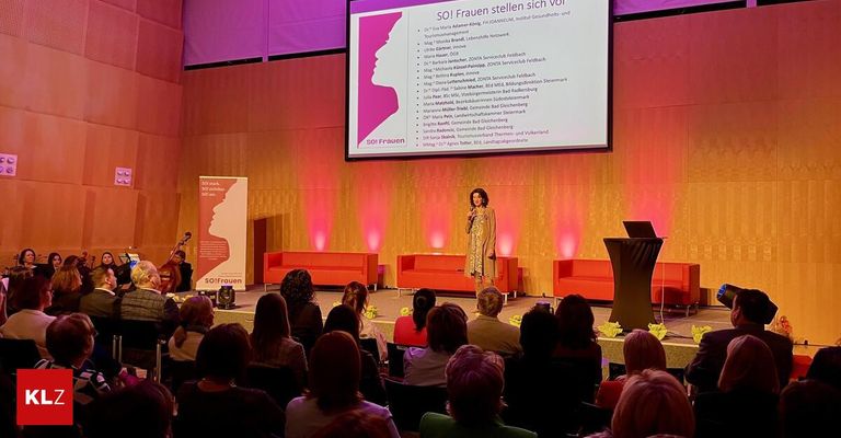 Eine Frau hält eine Rede auf der Bühne mit einer Projektionsleinwand hinter ihr. Die Leinwand zeigt eine Liste von Namen und Organisationen. Das Publikum sitzt in Stühlen vor der Bühne, und es gibt einen Rednerpult mit einem Laptop darauf.