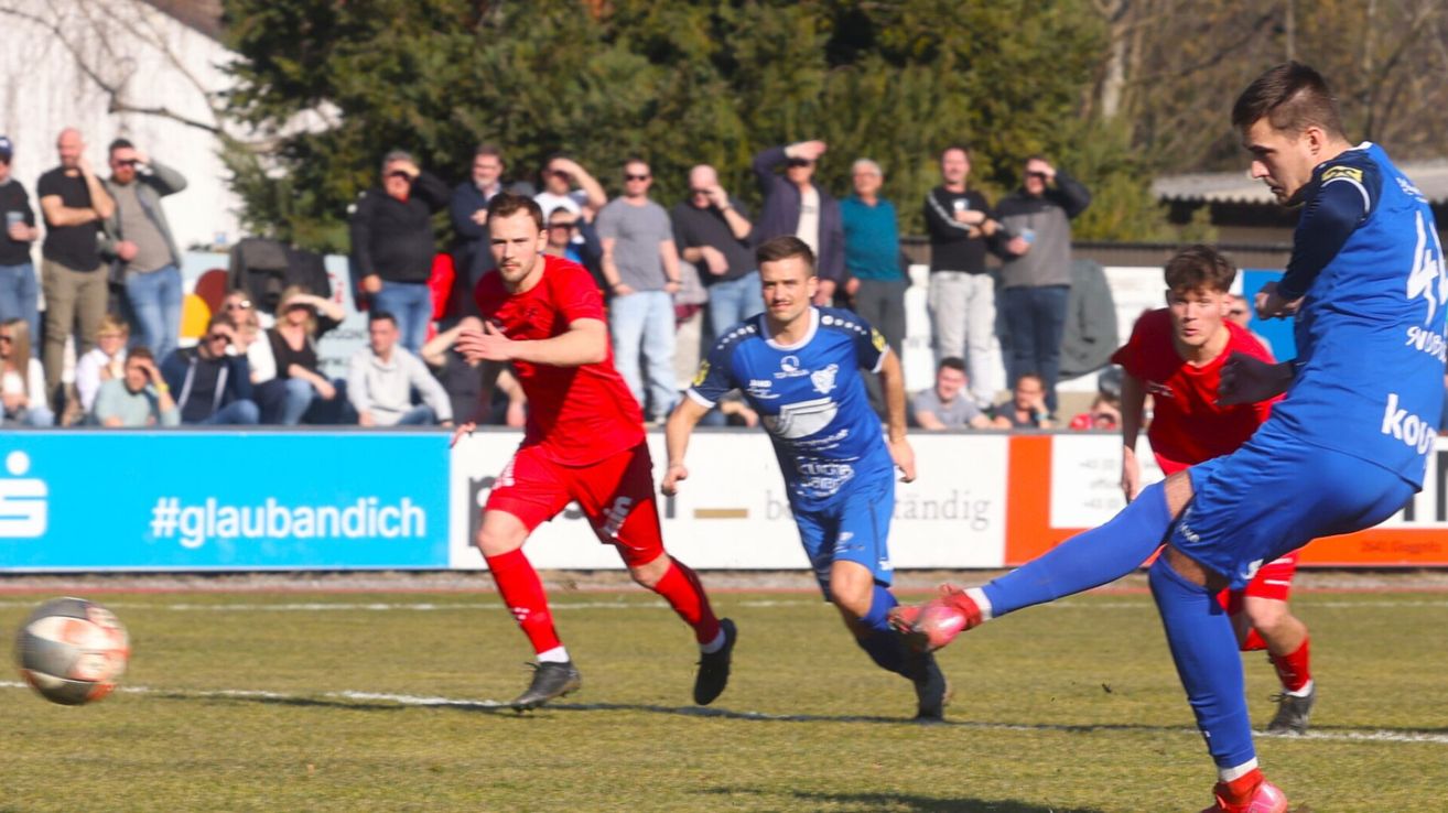 Zwei Fußballspieler in roten und blauen Trikots spielen auf dem Rasen, Zuschauer beobachten von einer Barriere aus.