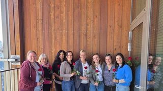 Eine Gruppe von Frauen posiert auf einem Balkon für ein Foto, jede hält eine rote Rose. Die hölzerne Wand hinter ihnen hat eine Tür und ein Glasfenster.