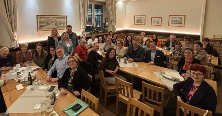 A group of people are seated around a table in a restaurant, smiling and posing for a photo. They are dressed casually, and some are wearing glasses. The table is set with plates, glasses, and napkins. Behind them are chairs and a window with curtains.