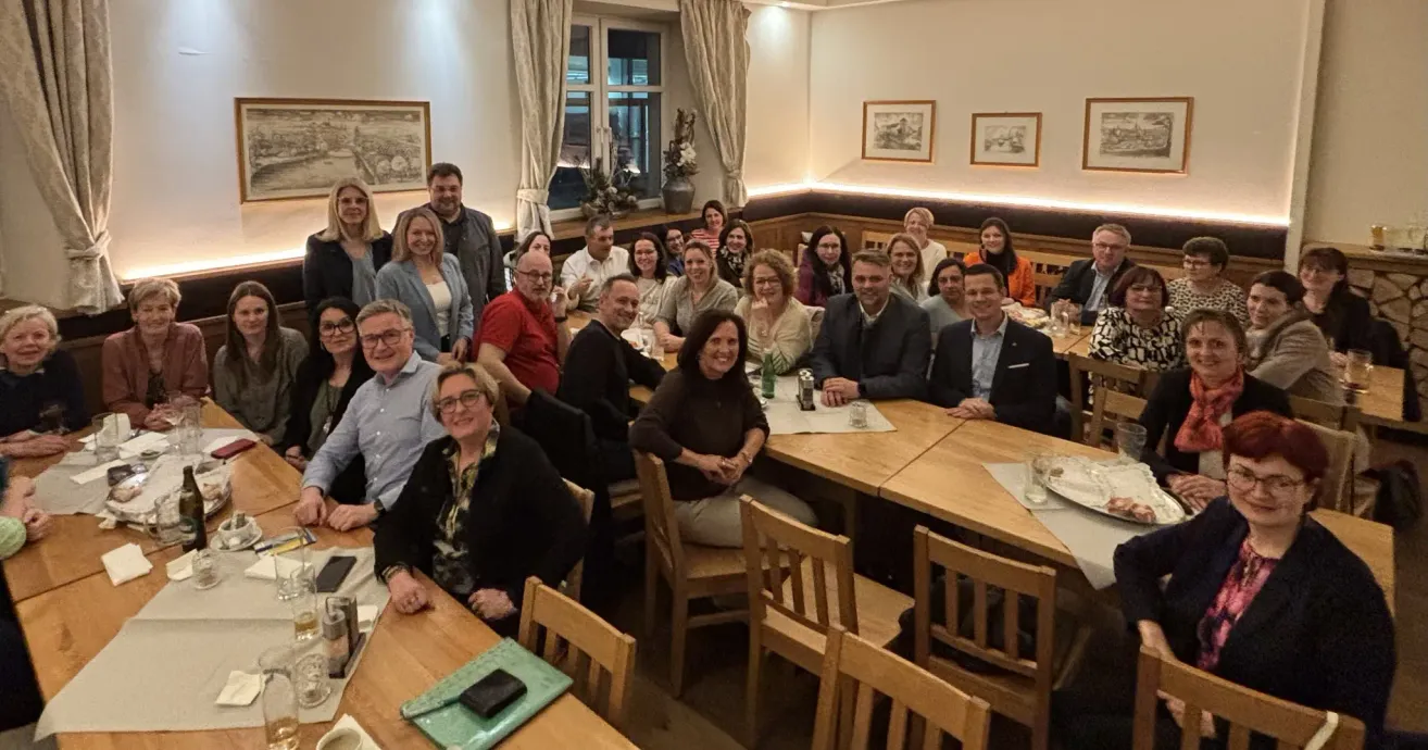 A group of people are seated around a table in a restaurant, smiling and posing for a photo. They are dressed casually, and some are wearing glasses. The table is set with plates, glasses, and napkins. Behind them are chairs and a window with curtains.