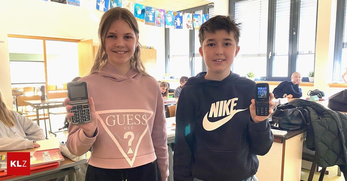 A boy and girl stand in a classroom, holding cell phones. The girl wears a pink sweatshirt with 'Guess' on it, while the boy wears a black Nike hoodie. Behind them, a classroom setting is visible with students sitting at desks.