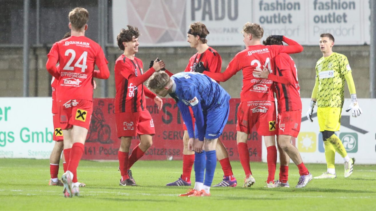 Fußballspieler in roten und blauen Trikots feiern einen Sieg auf einem verregneten Spielfeld, mit einem verschwommenen Werbebanner im Hintergrund.