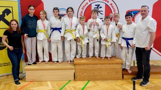 Eine Gruppe junger Kinder in weißen Kampfsportuniformen mit Medaillen um den Hals steht auf einem Podium. An der Wand hinter ihnen befindet sich ein Logo und der Text Europäische Judo-Föderation Österreich.