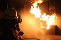 Ein Feuerwehrmann mit Helm und Maske löscht einen Brand, der einen Raum verschlungen hat.