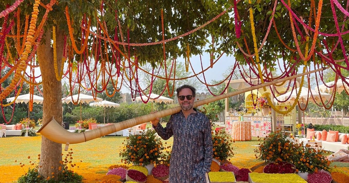 A man stands under a decorated tree holding a large horn. Colorful flowers surround him, and there are umbrellas and a festive table in the background.