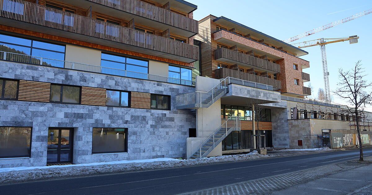 Modernes Gebäude mit Steinfassade, Glasfenstern, Holzbalkonen und einer Treppe zum Eingang. Der Boden ist schneebedeckt.
