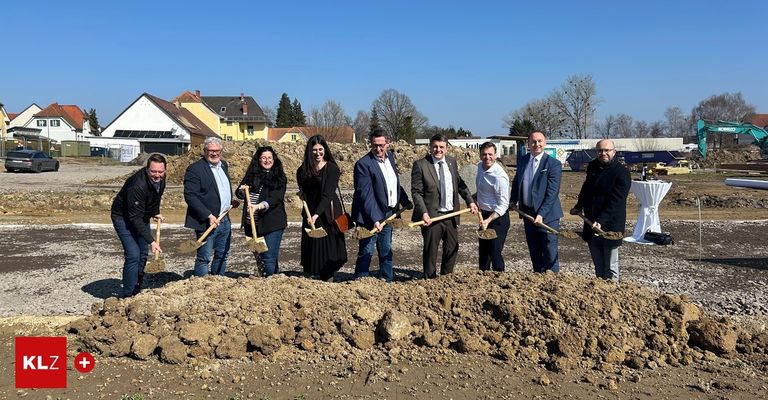 Group of people in business attire holding shovels at a construction site with houses and trees in the background.