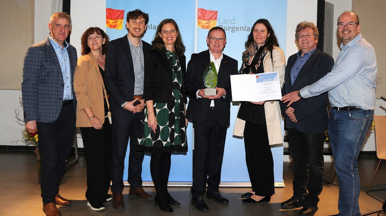 Eine Gruppe von Menschen steht zusammen vor einem blauen Hintergrund. Ein Mann in einem Anzug hält eine Trophäe und ein Zertifikat. Hinter ihm hält eine Frau ein Zertifikat. Die Gruppe lächelt und posiert für ein Foto.