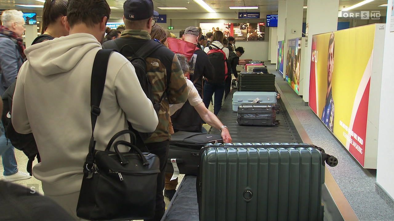 Reisende an einem Flughafen warten auf ihr Gepäck auf einem Förderband. Sie tragen verschiedene Jacken und haben Rucksäcke und Handtaschen bei sich.