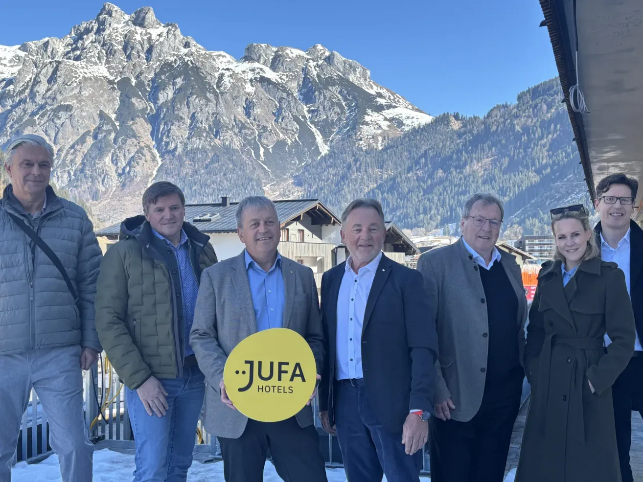Fünf Männer stehen vor verschneiten Bergen und lächeln. Einer hält einen gelben Kreis mit 'JUFA HOTELS'. Dahinter sind ein Haus und ein Schild zu sehen.