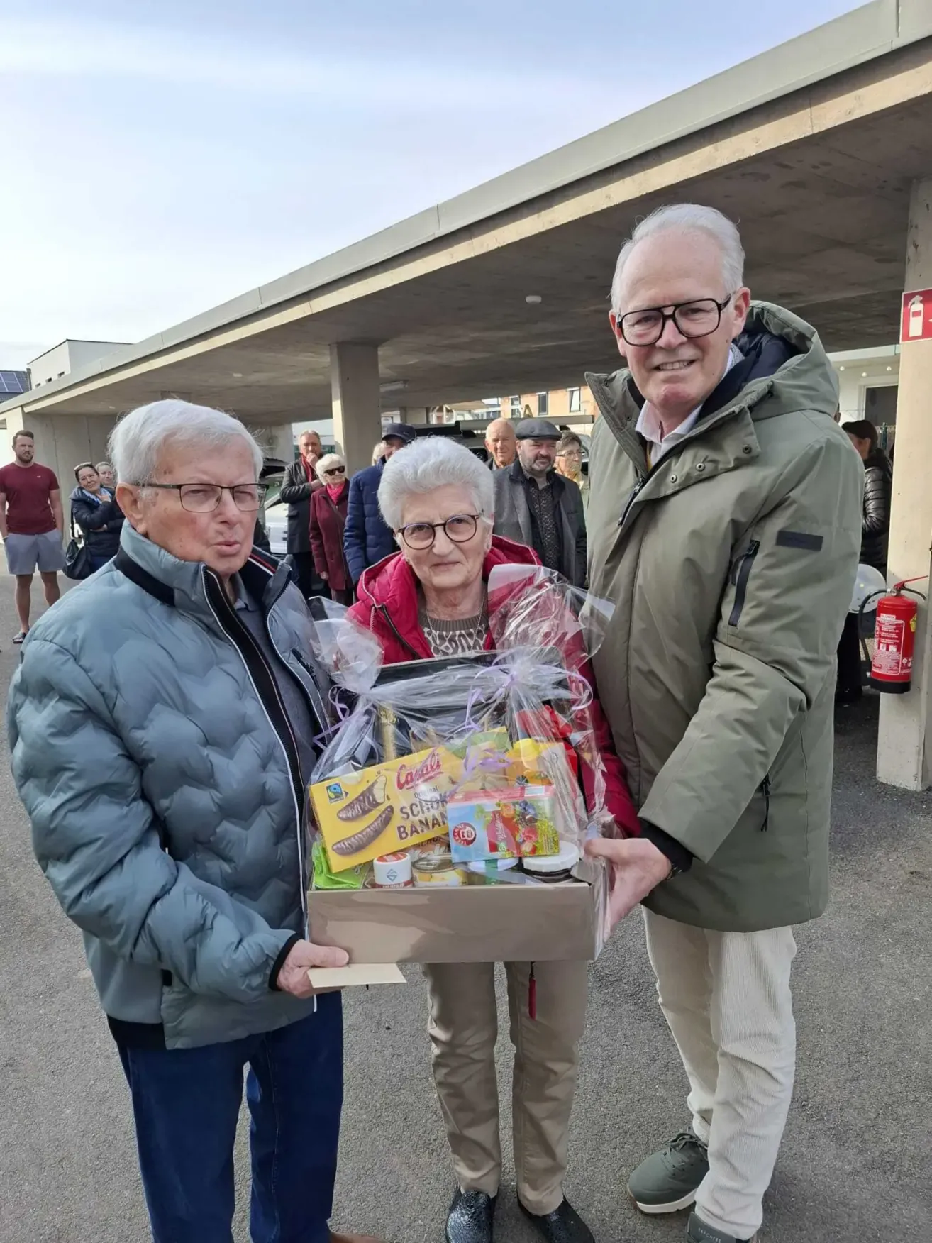 Three people are standing in a parking garage, one holding a box with a plastic cover. The elderly woman is wearing glasses, and the man beside her is smiling. Behind them, several people are standing, and some are looking at the car. A fire extinguisher is mounted on the wall.