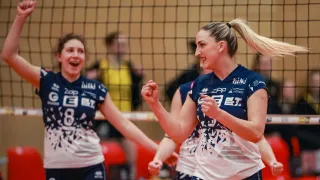 Three volleyball players celebrate on a court, one with a ponytail wearing a blue jersey with sponsor logos, while two others behind them are partially visible.