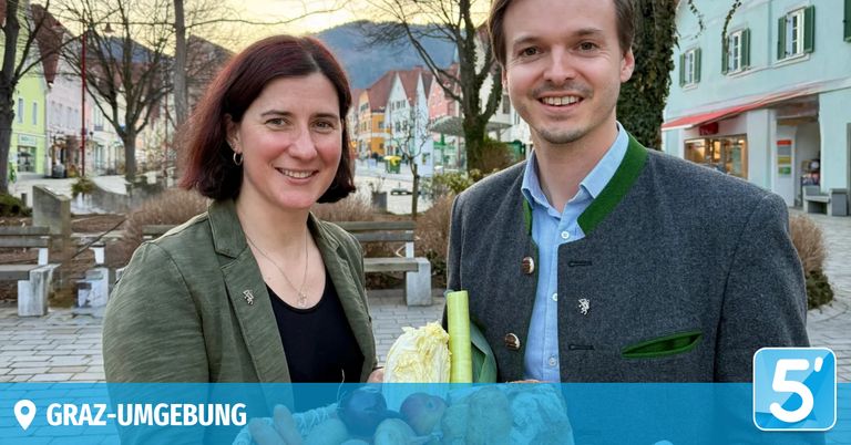A smiling man and woman stand in front of a picturesque town with mountains in the background. They hold a basket of vegetables.