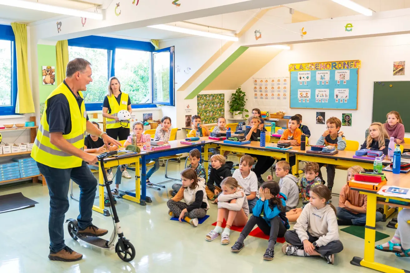 In a classroom, a man in a reflective vest is showing a scooter to children sitting around tables. A woman holds a helmet. A bulletin board and plants are in the background.