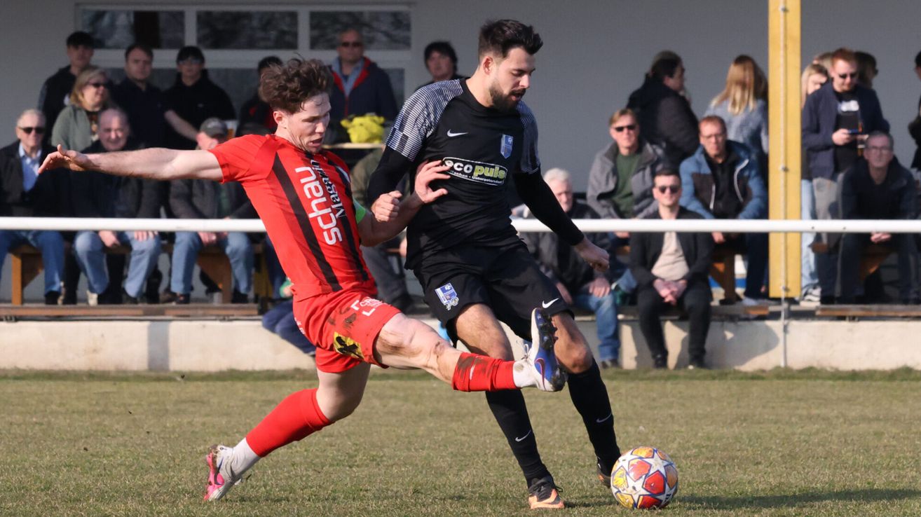 Two soccer players compete for the ball in an outdoor match. One player wears a red jersey with the word 'Schau', while the other wears a black jersey with 'Niko'. Spectators watch from the sidelines.