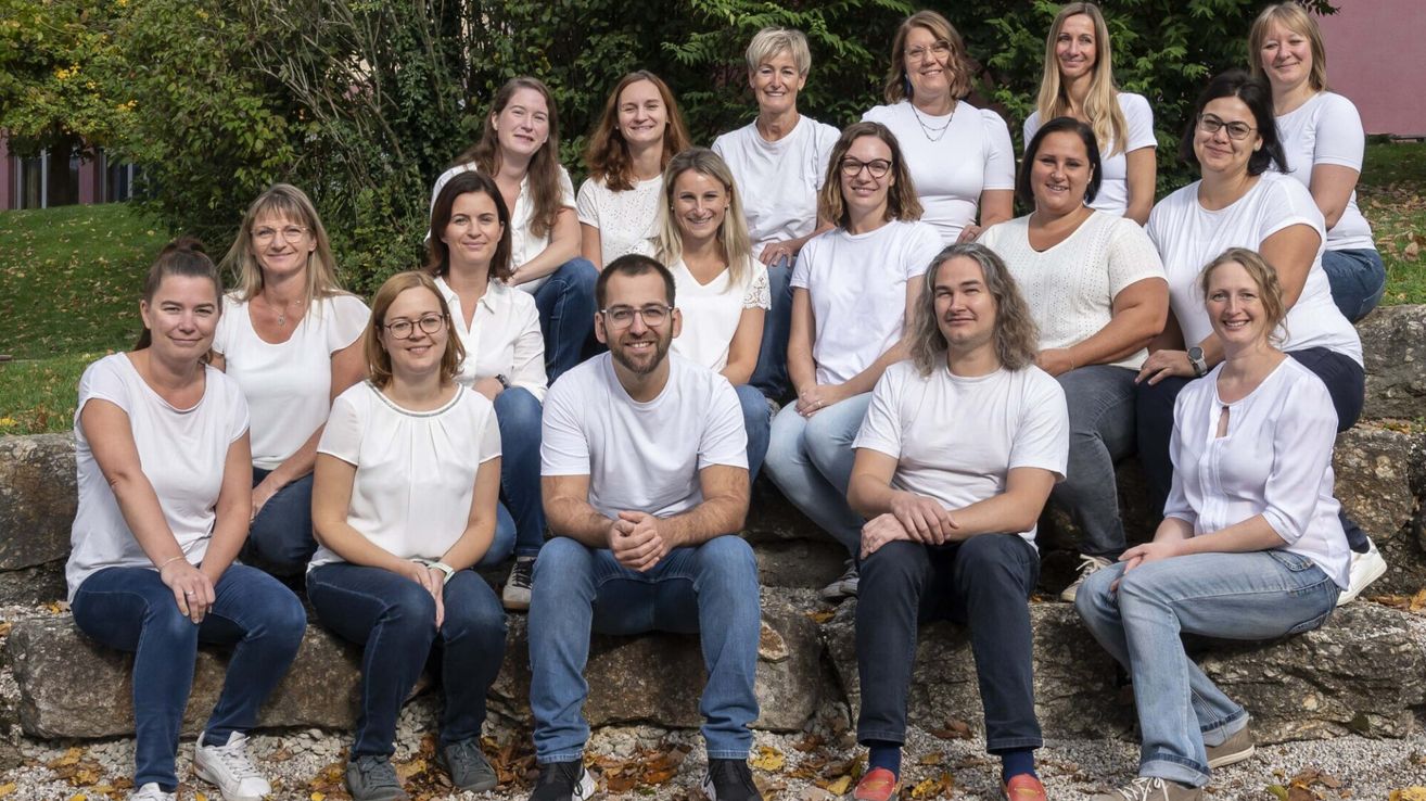 Eine Gruppe von Erwachsenen, sowohl Männer als auch Frauen, gekleidet in weißen Shirts, posieren für ein Foto im Freien. Sie sitzen auf Felsen, mit einer Kulisse aus Grün.