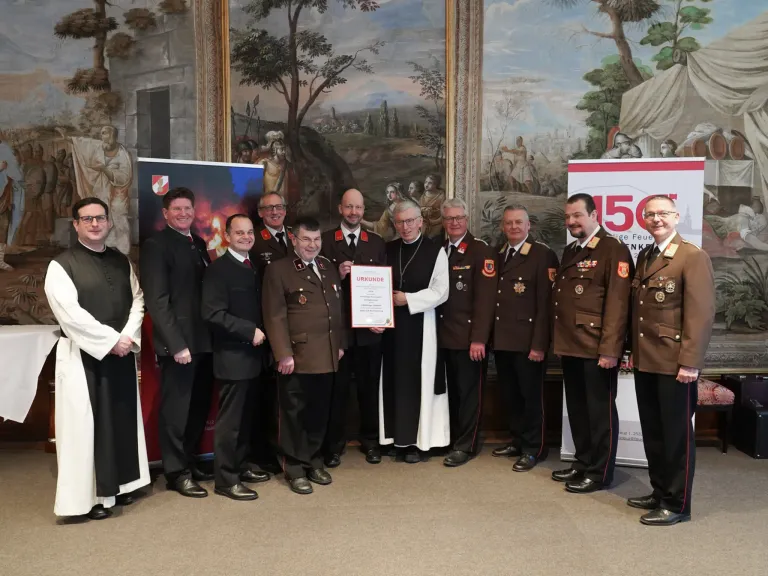 Eine Gruppe von Männern in Uniform und Anzügen steht vor einem Wandbild und erhält von einem Mann in einer weißen Robe eine Auszeichnung.