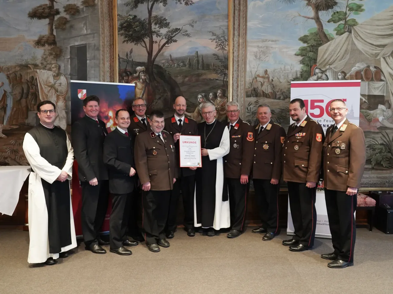 Eine Gruppe von Männern in Uniform und Anzügen steht vor einem Wandbild und erhält von einem Mann in einer weißen Robe eine Auszeichnung.
