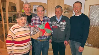 Five people stand in a room with a wooden cabinet. One man holds a wrapped gift, and another has a picture frame. The room has wooden flooring and a white wall.