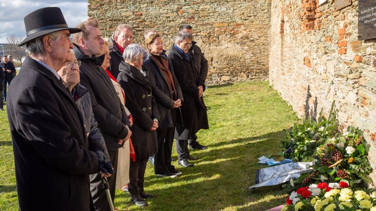 A group of people in winter coats stands in a solemn line in front of a brick wall, with some flowers and a folded flag nearby.