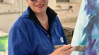 An elderly woman wearing a blue shirt and glasses stands in an art studio, smiling while painting a canvas with a brush. Behind her is a partially visible wall with a window.