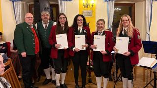Five people stand together holding certificates in a room with a yellow wall and a chandelier. They wear red jackets and black pants, except for the man on the left in a green vest.