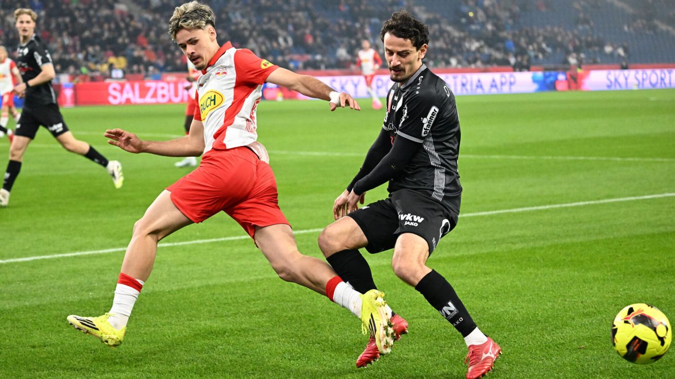 Two soccer players in action on a field. One player in red shorts and a white jersey, the other in black with a yellow logo.