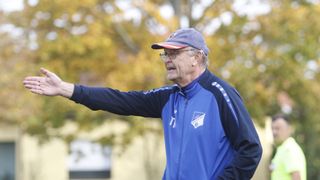 An older man with a cap and glasses is standing and appears to be giving instructions, pointing his arm outward. He is wearing a blue jacket with a logo and the word 'Haltern'. Behind him, there are blurry trees with yellow leaves.