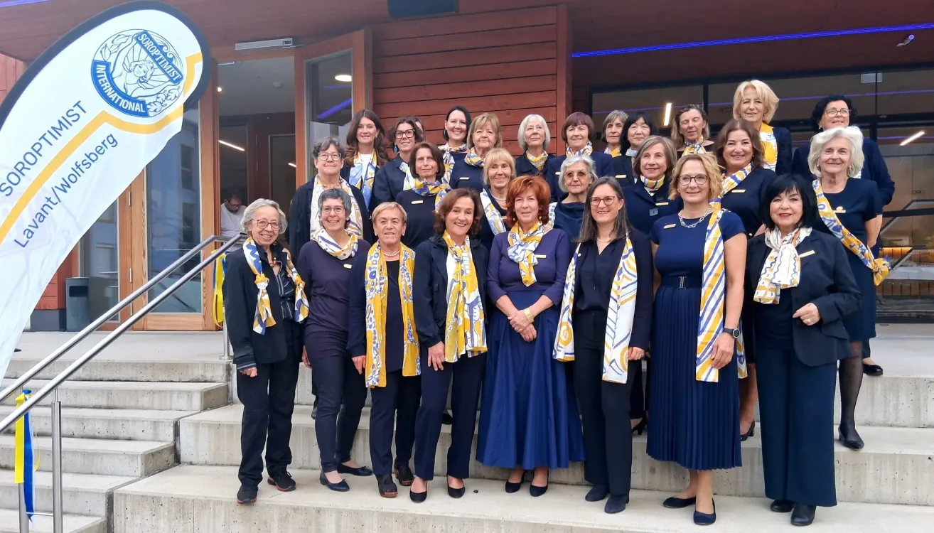 A group of women, dressed in matching uniforms, stand on the steps of a building, smiling for a photograph. They are all wearing yellow and white scarves.