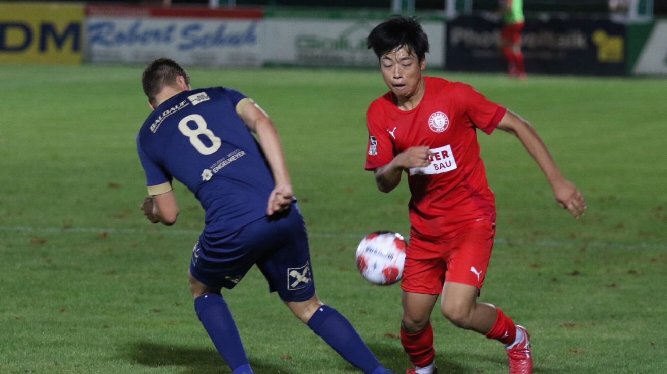 Two soccer players compete on a field; one in a blue jersey with number 8, the other in a red jersey with text 'GER BAU', chasing a soccer ball.