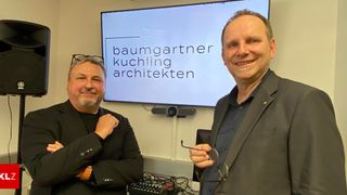 Zwei Männer stehen lächelnd vor einer Beamerleinwand mit dem Namen Baumgartner Kuchling Architekten.