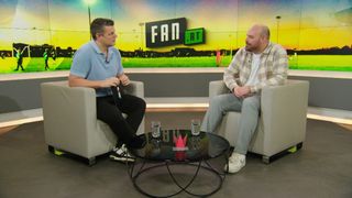 Two men sit on chairs, one holding a book, in front of a table with glasses. A large screen in the background shows a soccer field and the word Fan.