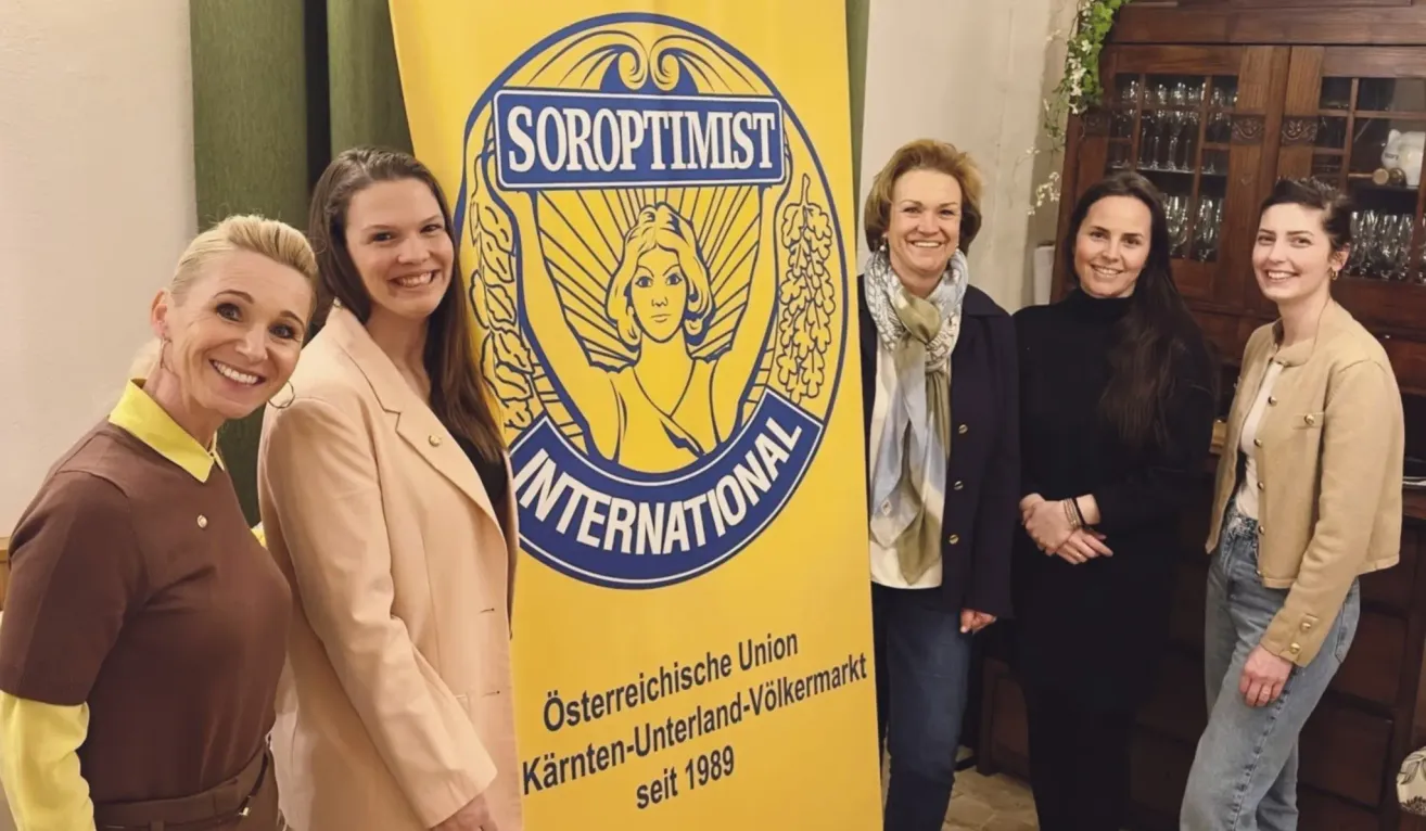Four women stand smiling in front of a yellow banner that reads 'Soroptimist International' with a logo of a woman holding wheat. The banner also says 'Austrian Union Karnten-Underland-Volkermarkt since 1989.'