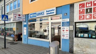 The storefront of Weiss Service, with a blue wall, a glass door, and a sign reading 'Weiss Service'. A trash bin is in front.