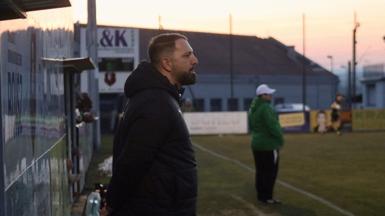 A man in a black hoodie stands on a sports field, watching another person in a green jacket and white cap. Behind them, a building with a sign reading '&K' is visible.