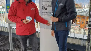 Two men in winter jackets stand near a donation box with a sign that reads Caritas & Du. They are on a balcony with mountains in the background.